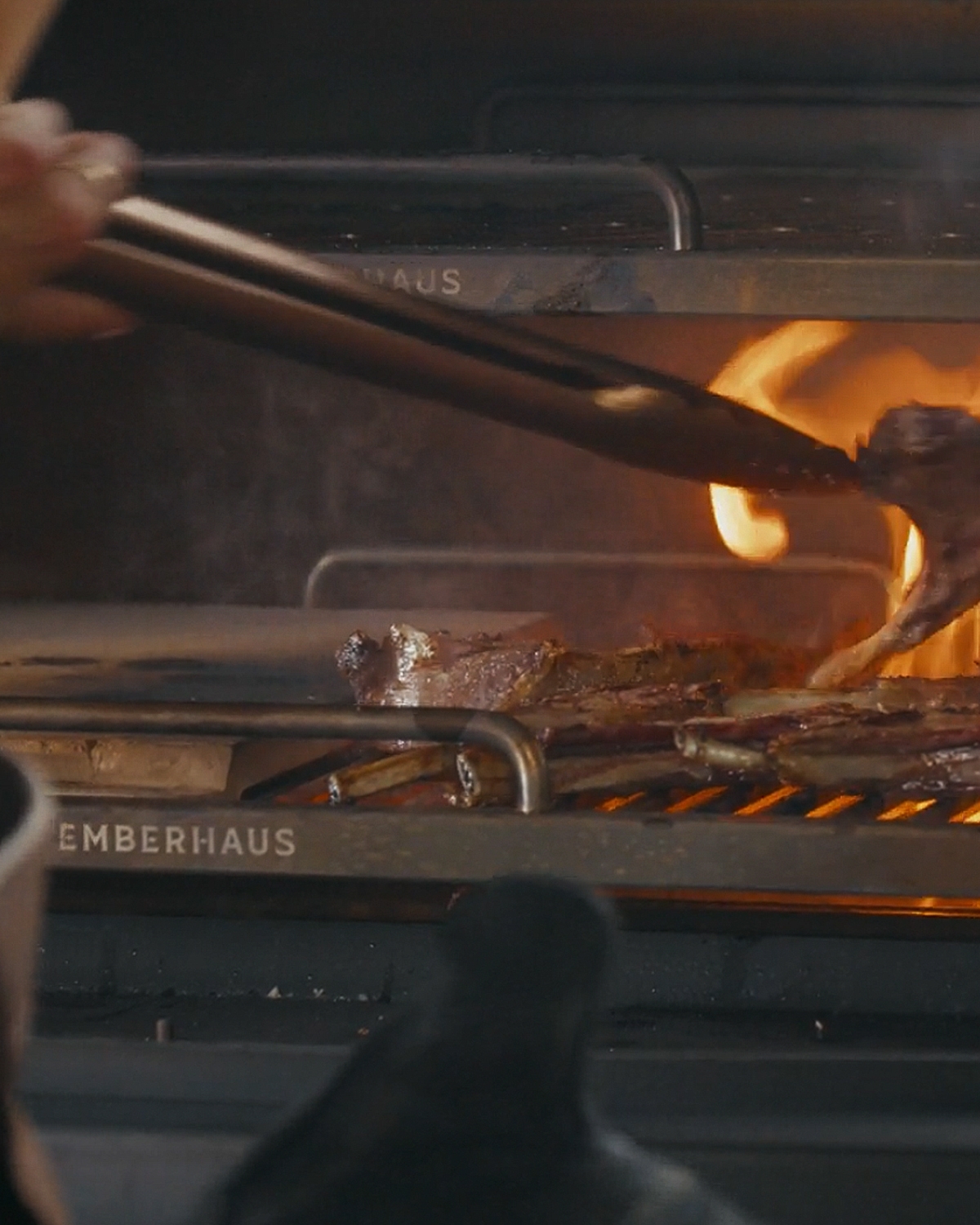 Close-up of an Emberhaus Fire Oven in action with split cooking surfaces, as flames rise beneath sizzling lamb chops, with a chef using tongs to tend the food—capturing the intensity and precision of live-fire cooking.