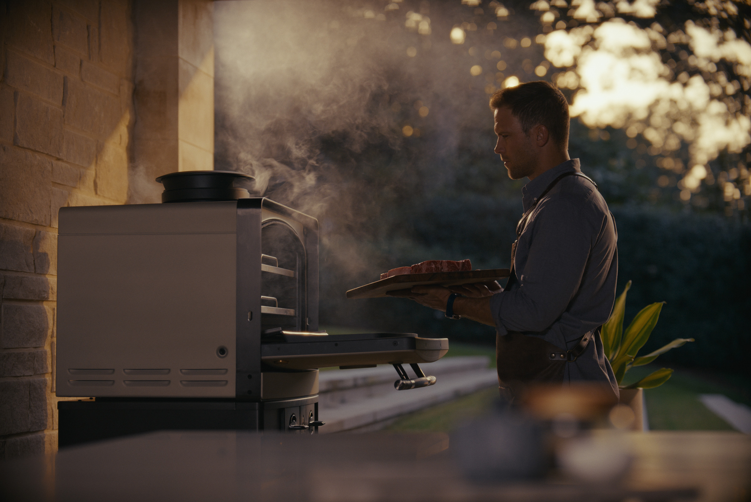 Man placing steaks into an Emberhaus Fire Oven at dusk, with smoke rising from the flame, showcasing live-fire cooking and luxury outdoor kitchen design.