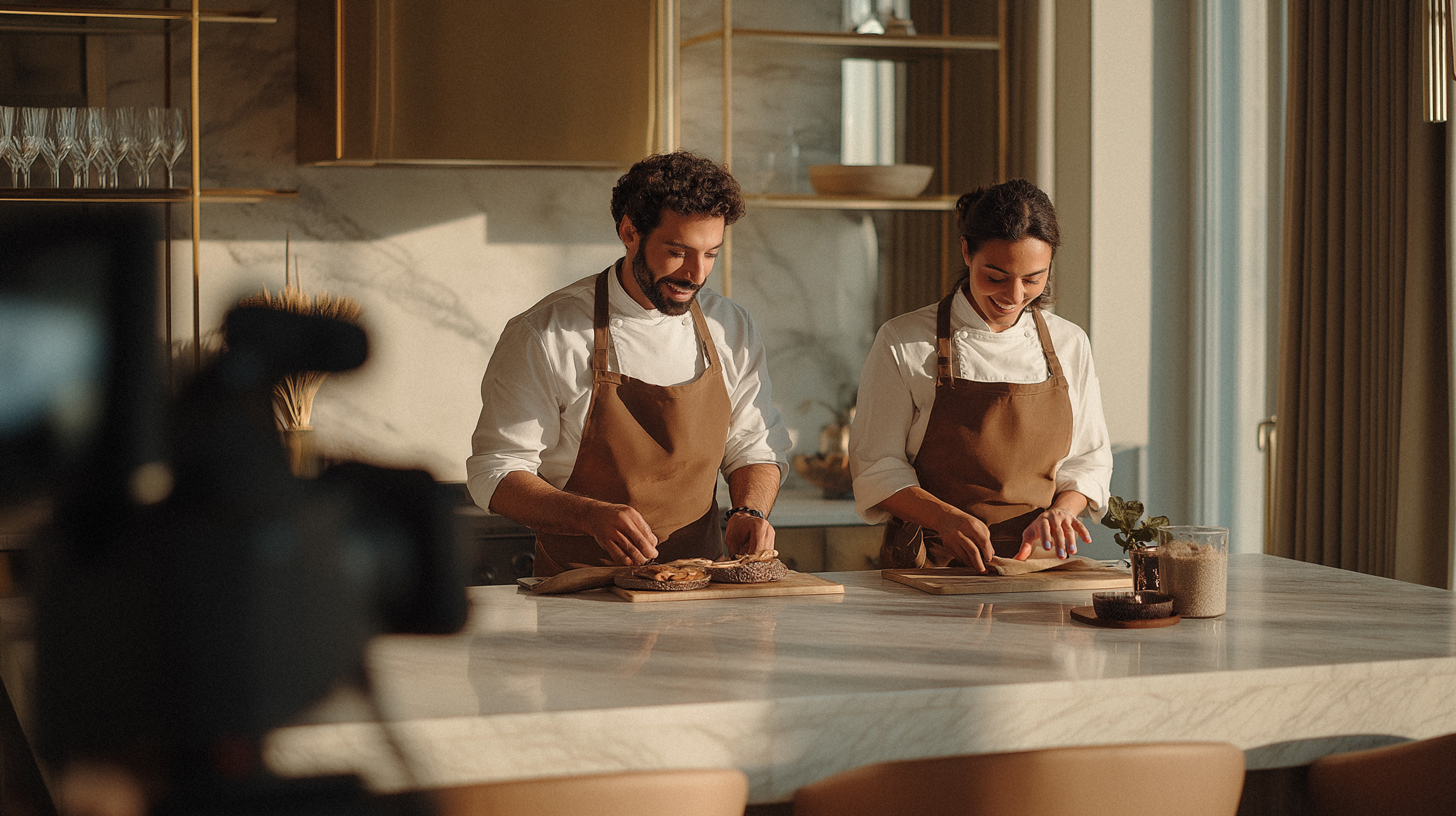 Two Emberhaus chefs at a kitchen island preparing a meal with a camera in the foreground filming.