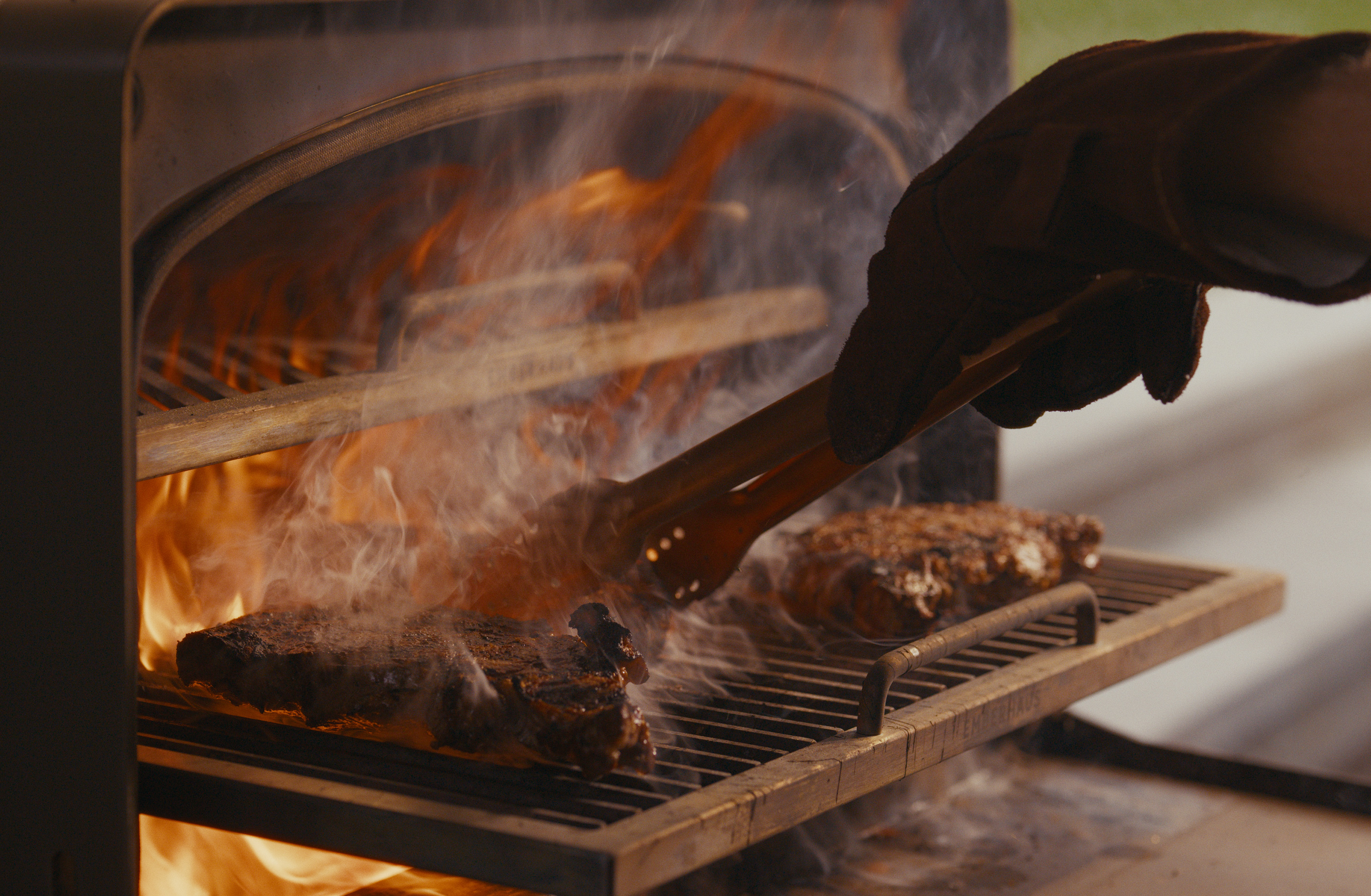 Grilling steak on Emberhaus Fire Oven, roaring flames, tongs.