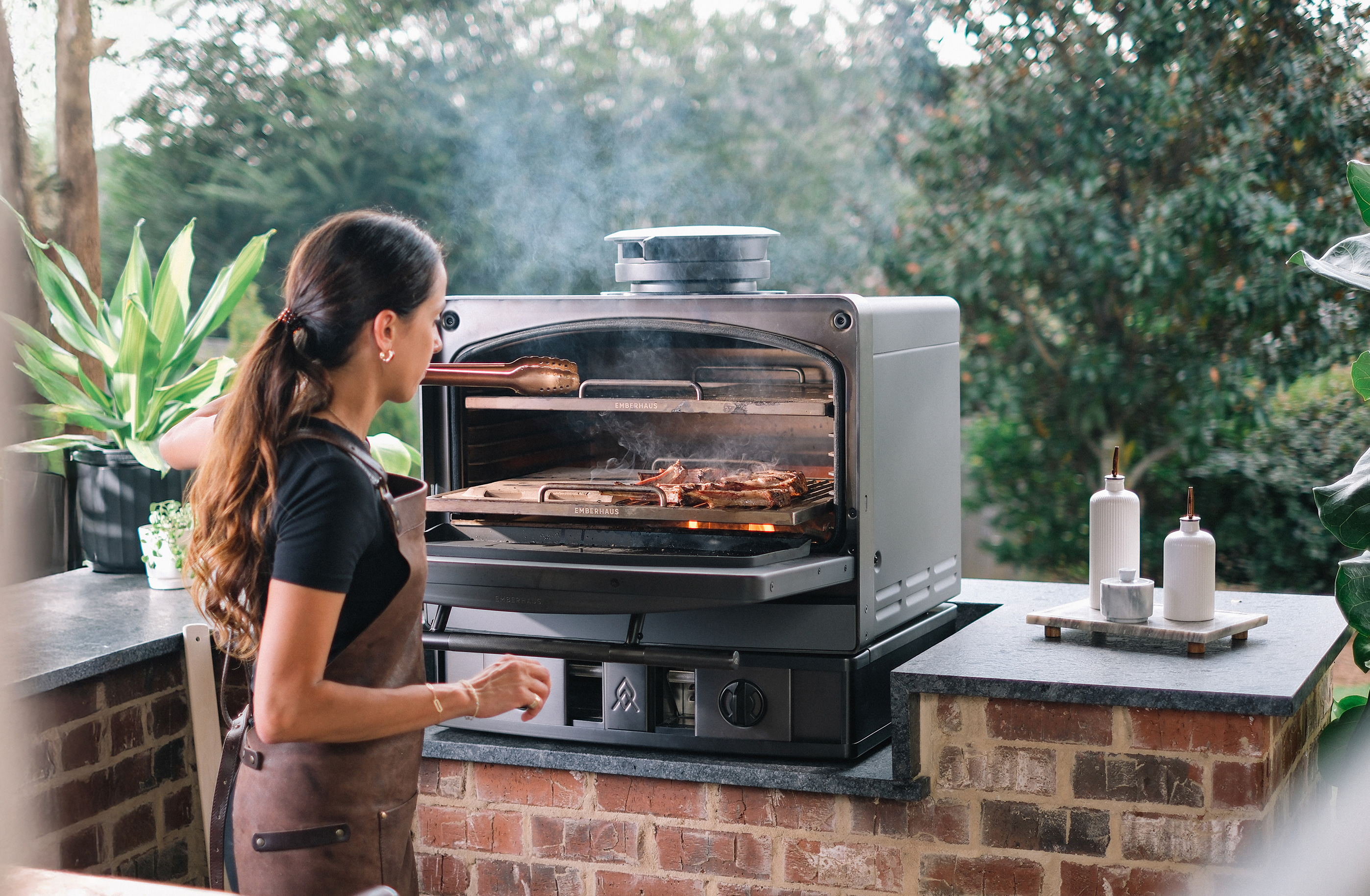 Woman cooking over an open flame in an Emberhaus Fire Oven, showcasing live-fire cooking, craftsmanship, and luxury outdoor kitchen design.