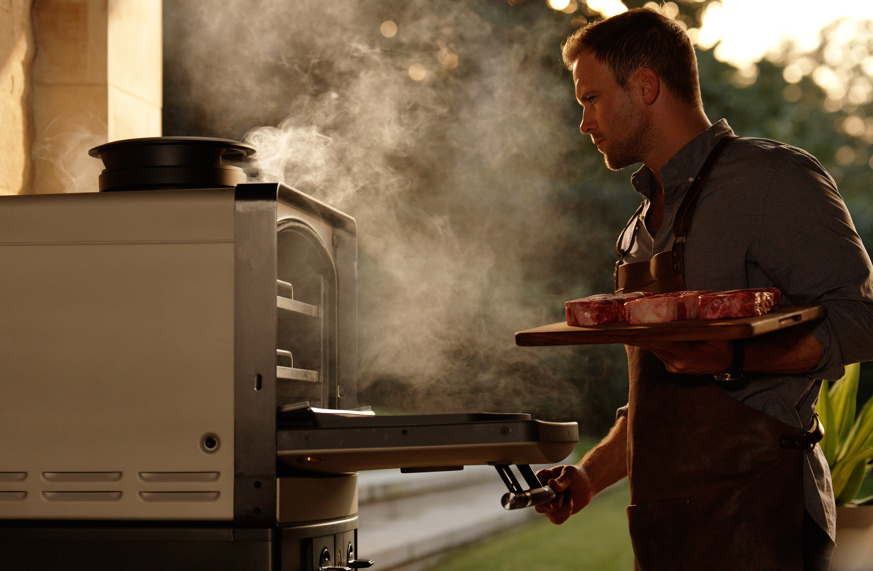 Man preparing steaks beside an Emberhaus Fire Oven, surrounded by smoke and warm light, showcasing live-fire cooking and luxury outdoor kitchen design.